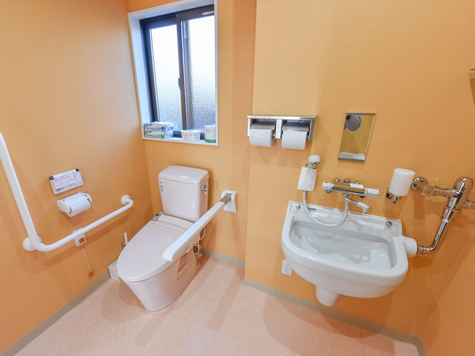 トイレの壁は、明るい黄橙色のデザインになっている。左端には窓があり、その手前に予備のトイレットペーパー等が置いてある。