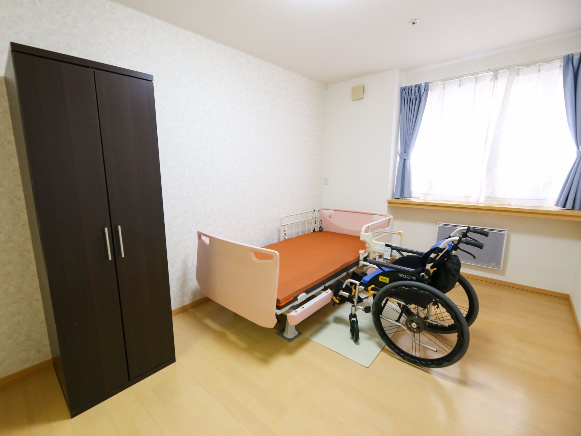 クローゼットや介護ベッド、ヒーターが完備された居室である。ベッドサイドに車椅子が置かれている。自然光が差し込む窓が設けられている。