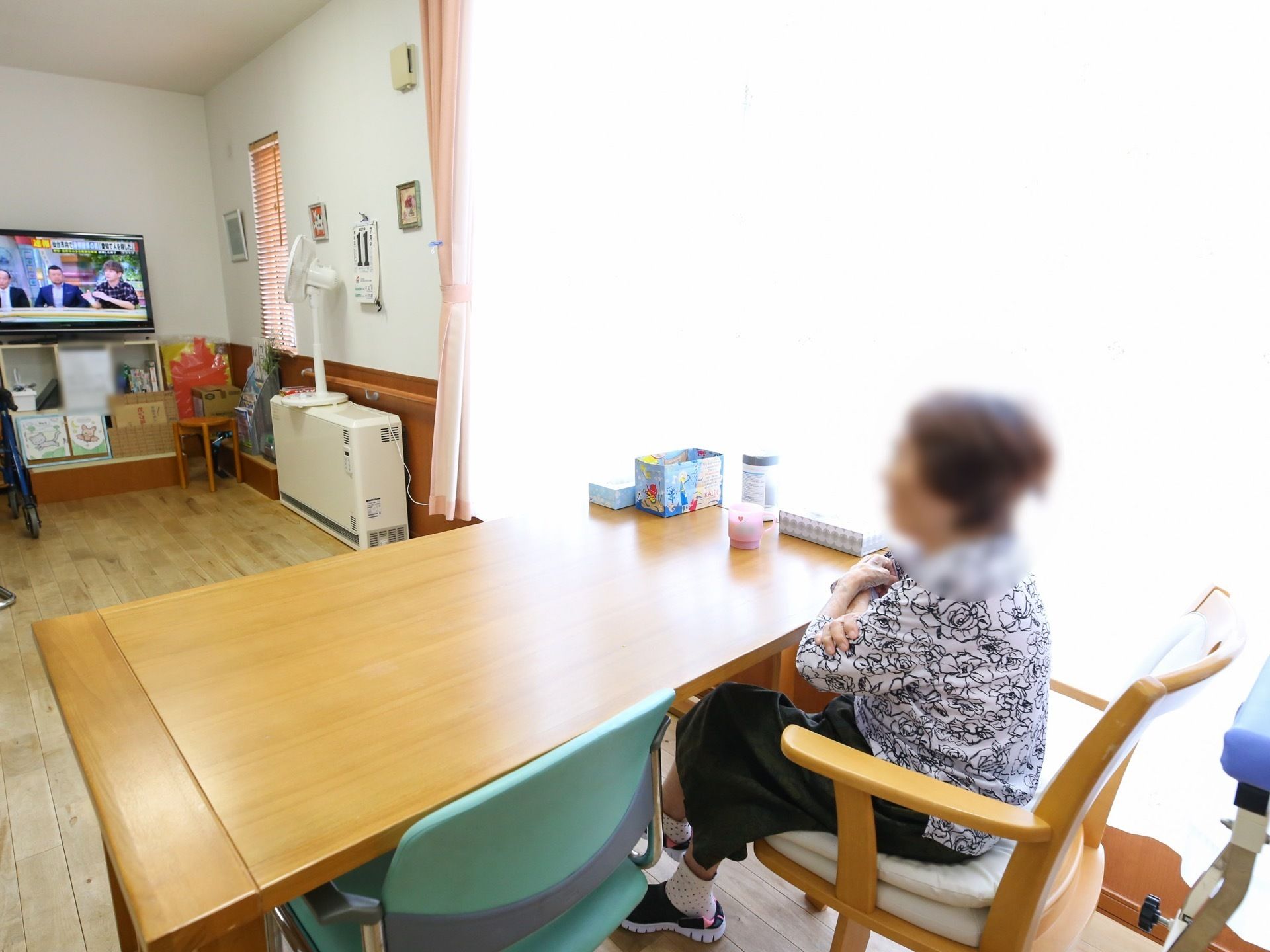 明るいダイニングルームである。窓際のテーブル席に座って入居者様がテレビを見ている。壁には、手すりが取り付けられている。