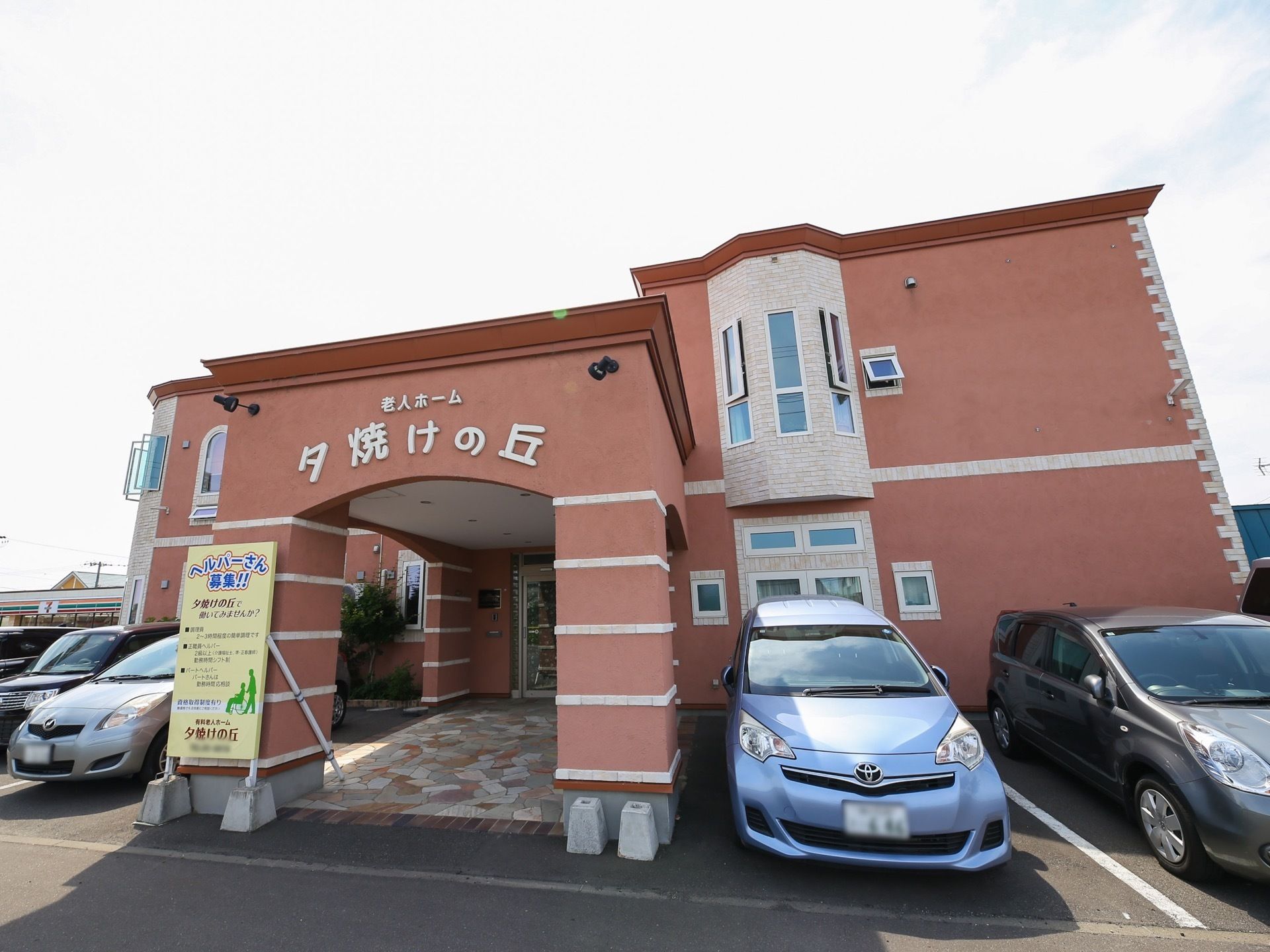 赤い塗り壁の２階建ての建物の周囲に、複数の自動車が駐車してある。玄関の前に屋根がアーチ状に伸びており、正面に施設名称が明示されている。