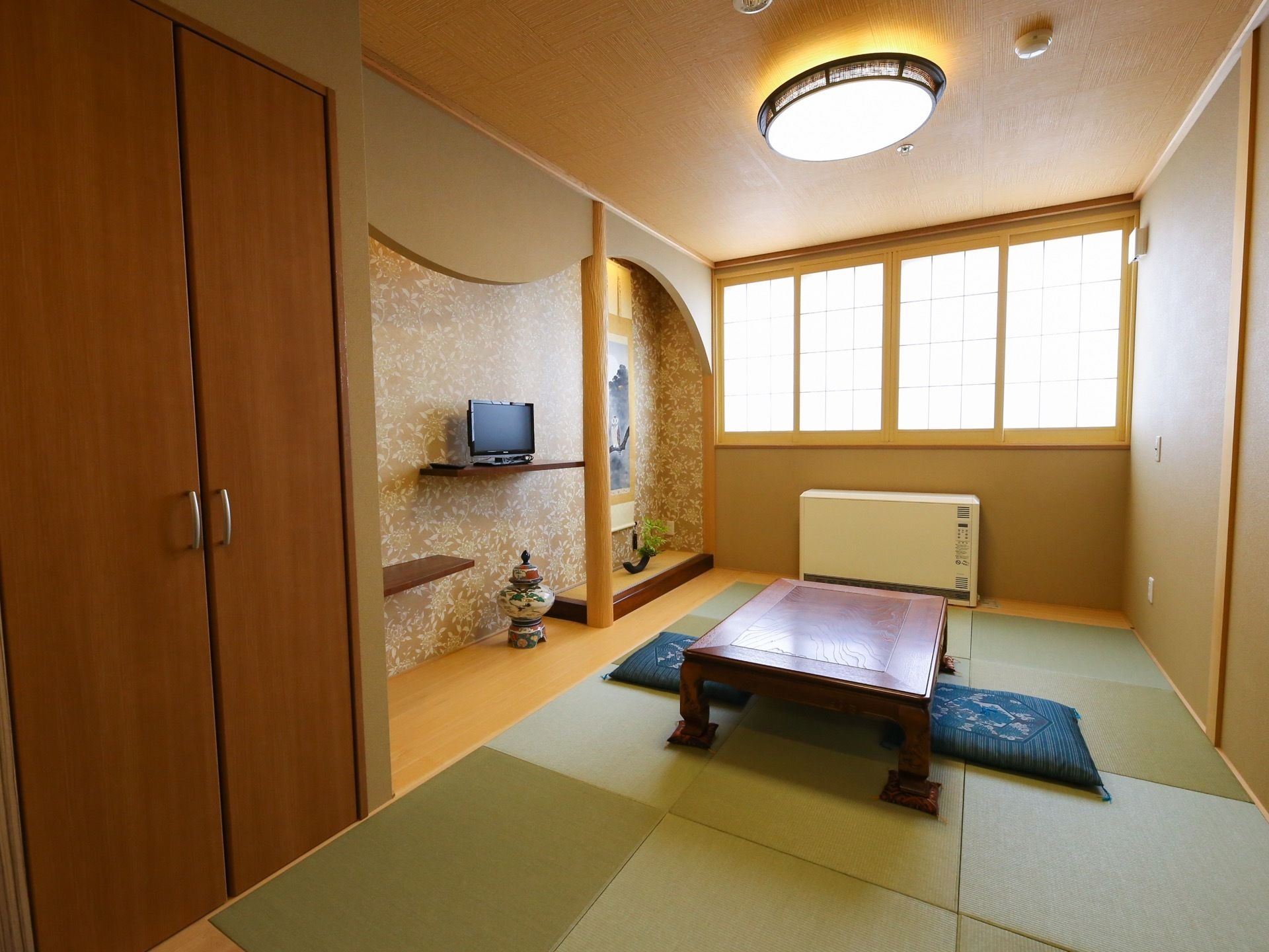 窓辺に障子戸が取り付けられた部屋に置き畳が敷き詰められ、座卓や座布団が配置されている。床の間に掛け軸や壺、液晶モニターがある。