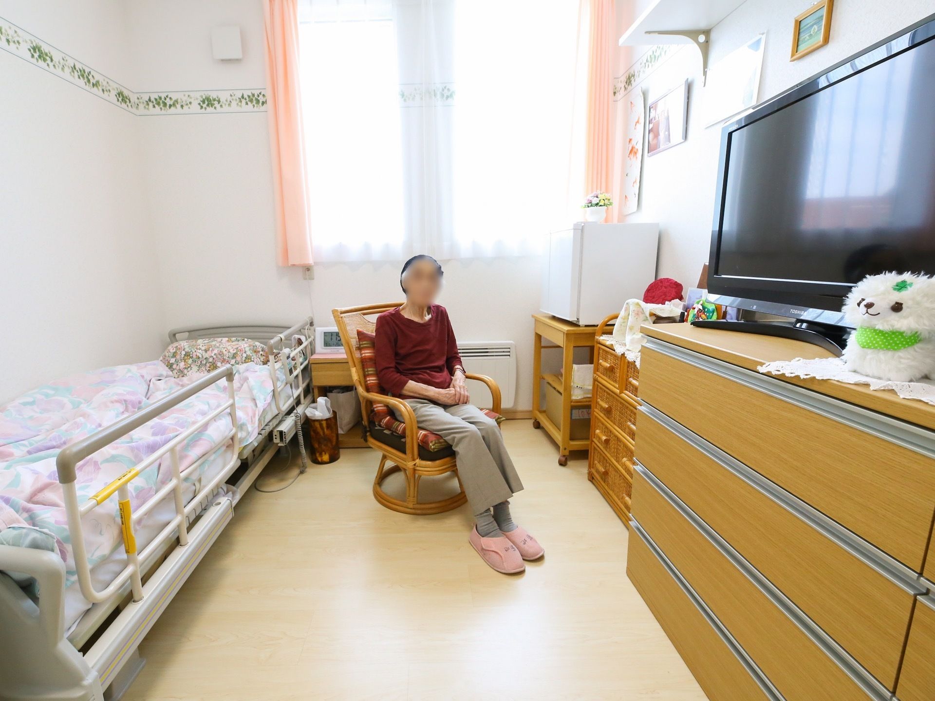 窓のある明るい室内に、ベッドやチェストなどの大きな家具が配置されている。部屋の中央に、椅子に座っている人物が見える。