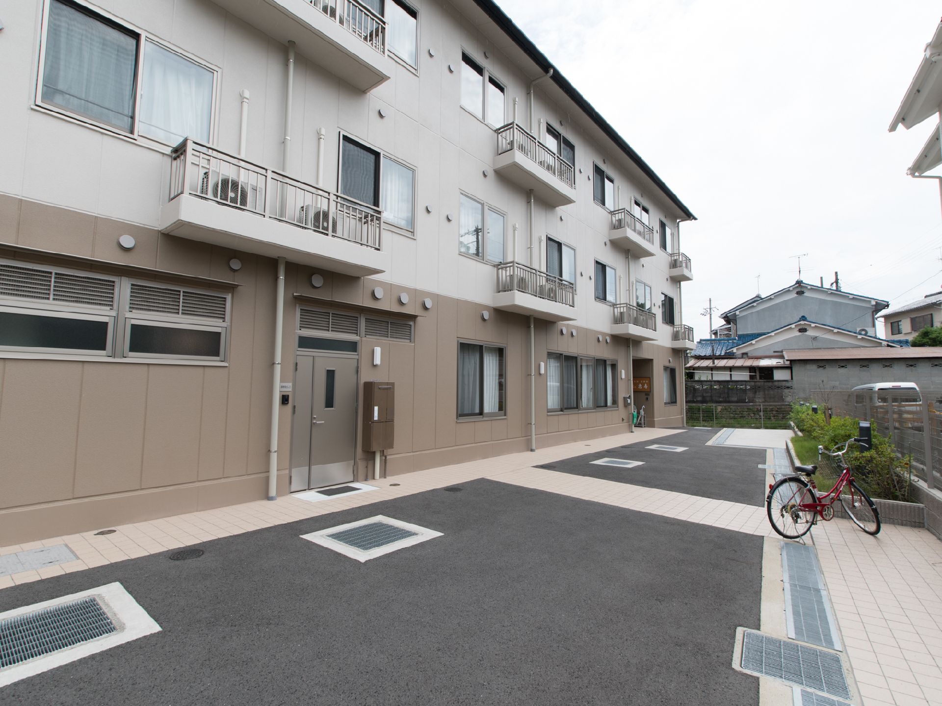 暗いベージュと明るめのベージュで塗られた3階建ての建物で、建物名が記載される看板の近くには傘立てが設置されている。