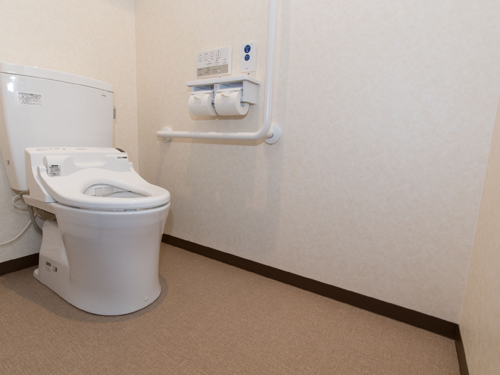 白を基調としている清潔感のあるトイレである。温水洗浄機能が付いていて、操作パネルが壁にある。手すりを完備している。