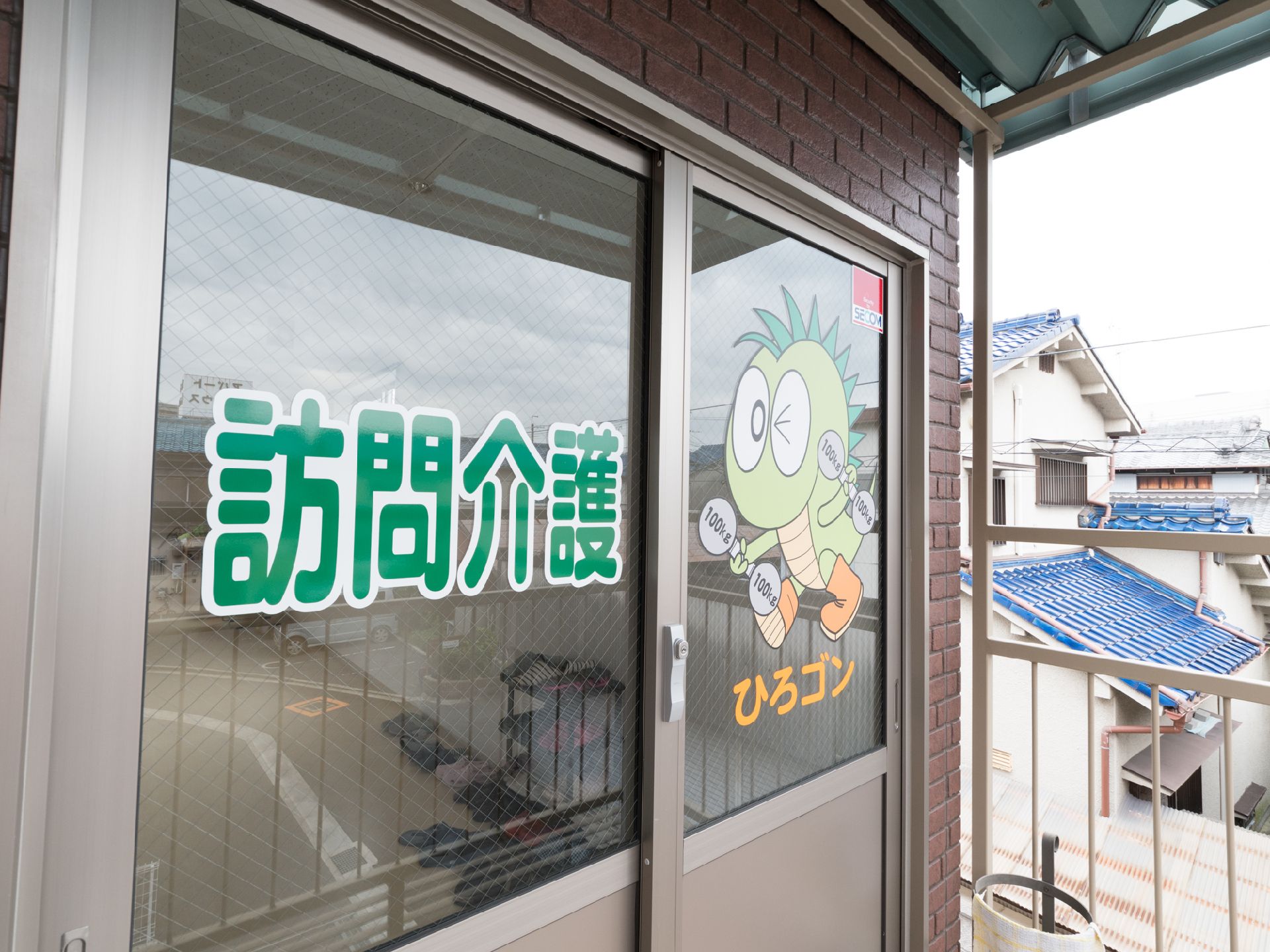 施設の向かいの建物２階には、訪問介護の事務所が入っている。傘立てが庇付きの玄関アプローチに置かれている。