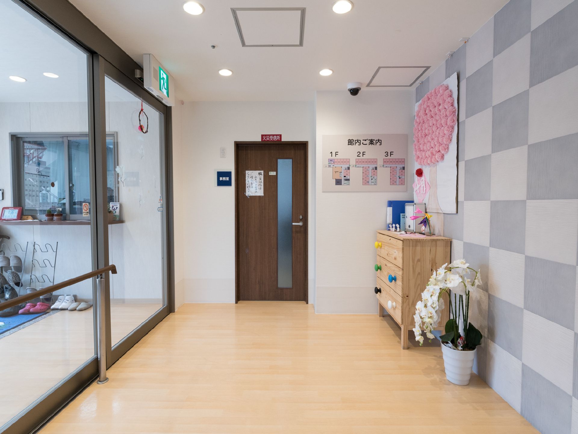 広いエントランスには、収納家具や胡蝶蘭が置かれている。館内の案内図が壁に設置されている。自動ドア近くに事務室の扉がある。