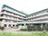 ５階建ての施設で、周囲は柵で囲まれている。入口の門は開けられ、門の横に掲示板がある。車が１台停まっている。