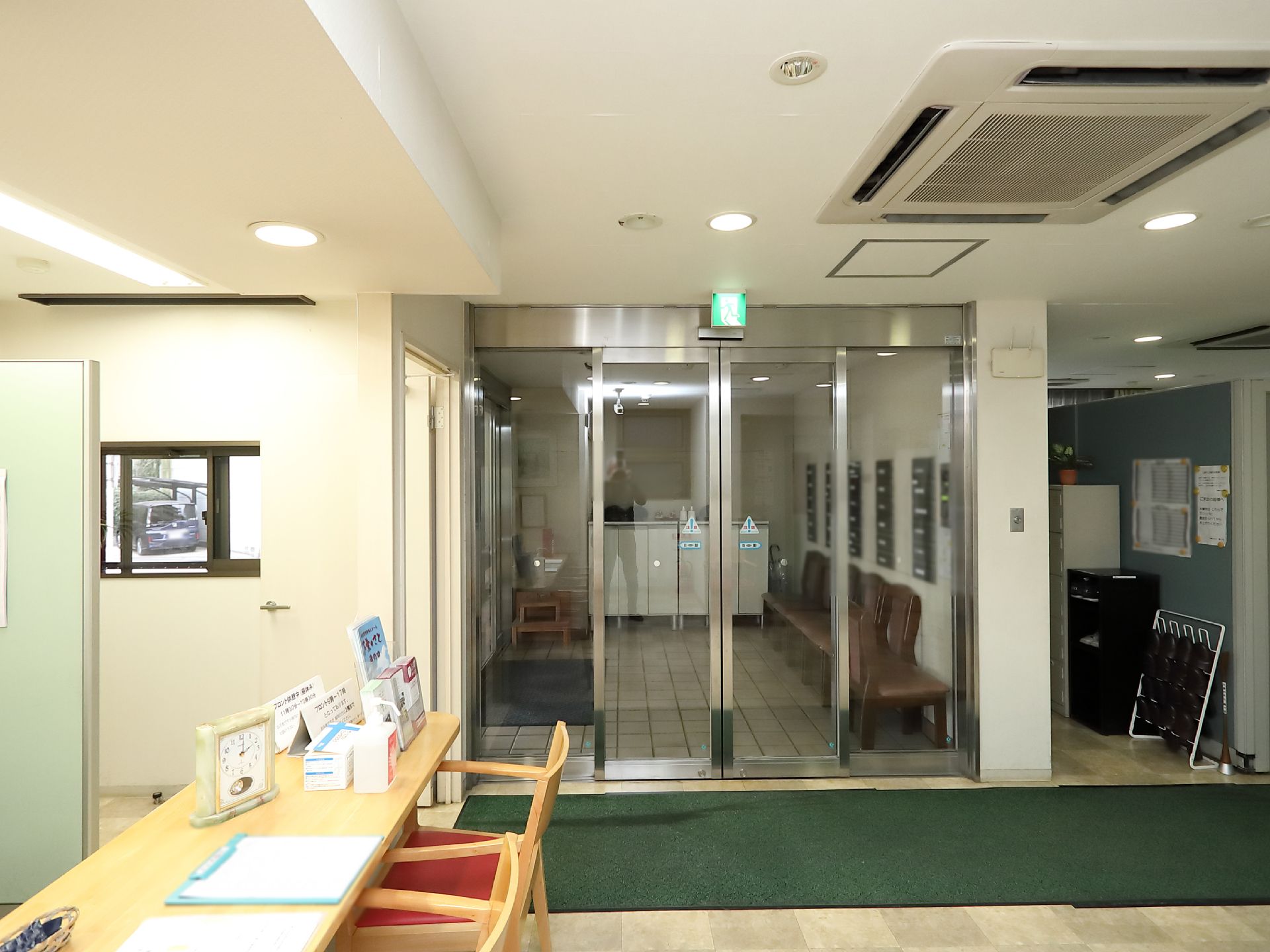 自動ドアの入り口で、壁にポストが設置されている。ポストの下に椅子が用意されている。床に緑の大きな玄関マットが敷かれている。
