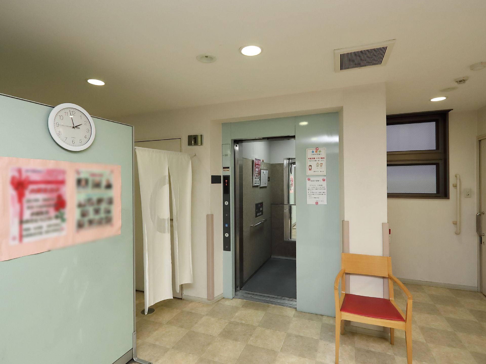 薄いブルー色ドアのエレベーターが設置されている。入り口付近の柱には衝撃緩衝材が取りつけてある。柱の前に椅子が置かれている。