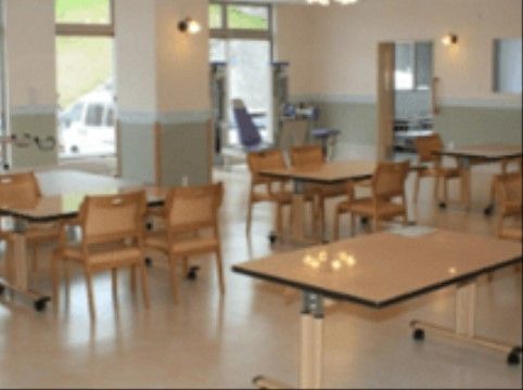 キャスター付きのテーブルとひじ掛け付きの椅子が複数置かれている。部屋の角にリハビリ器具が設置されている。