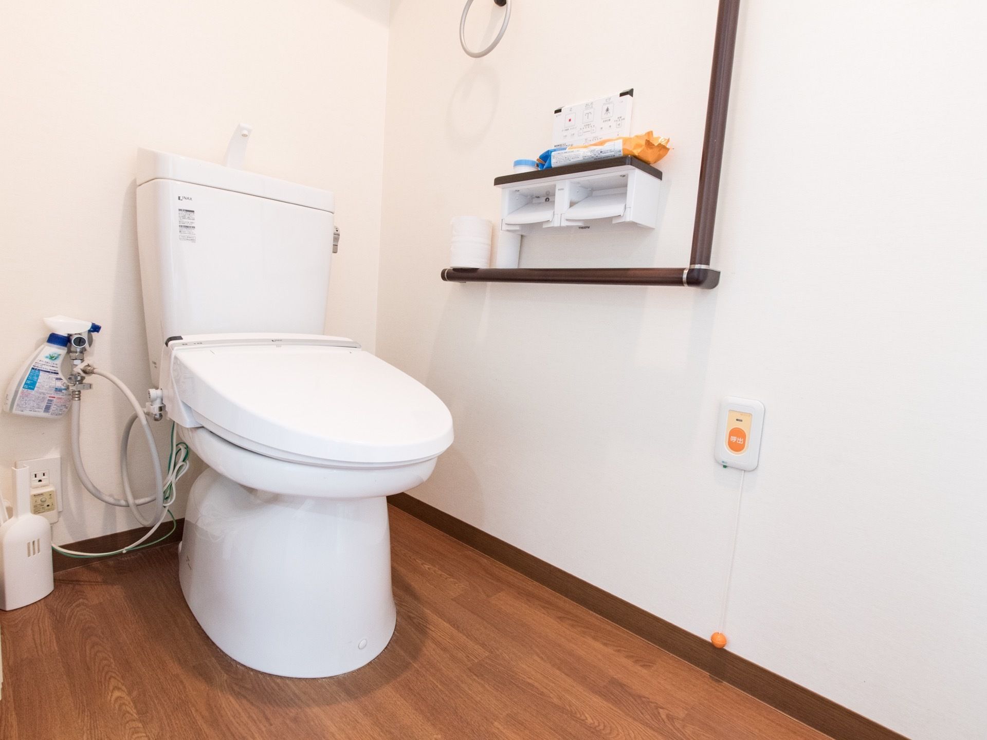 トイレは段差のないフローリングである。手すりや緊急ブザー、タオルハンガーが壁に取りつけられている。清掃が行き届いている。