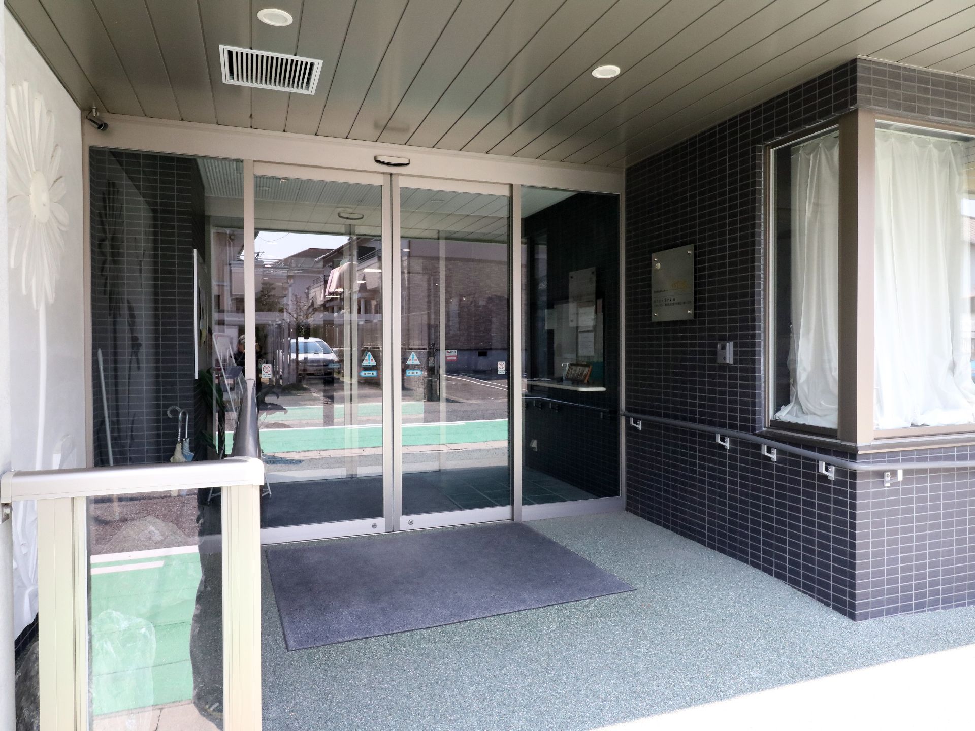ガラス製の自動扉のついた玄関。雨天の時など汚れを落としたりする玄関マットが敷いてある。車椅子でも行き交えることが出来る、十分な広さである。