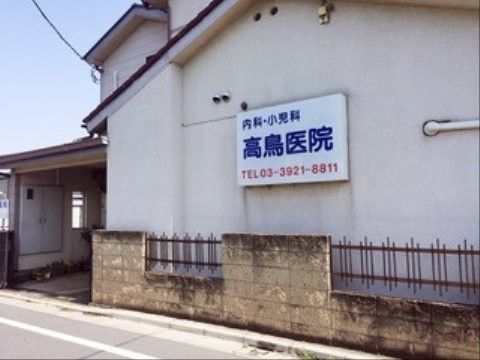 白い壁の建物の壁に、医院の看板が掛かっている。白地に青色の文字で院名が書かれ、赤色の文字で電話番号が書かれている。