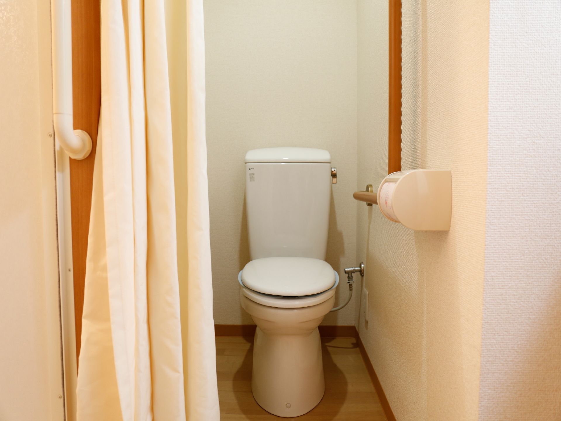 ナチュラル系の床と白いクロスが明るい雰囲気のトイレ。手すり、目隠し用のカーテンが取り付けられている。