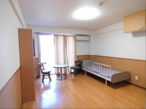 フローリング張りで、白壁に腰板の付いた室内に、電動介護ベッドや小さなテーブルと椅子、棚が置かれている。エアコンが設置されている。