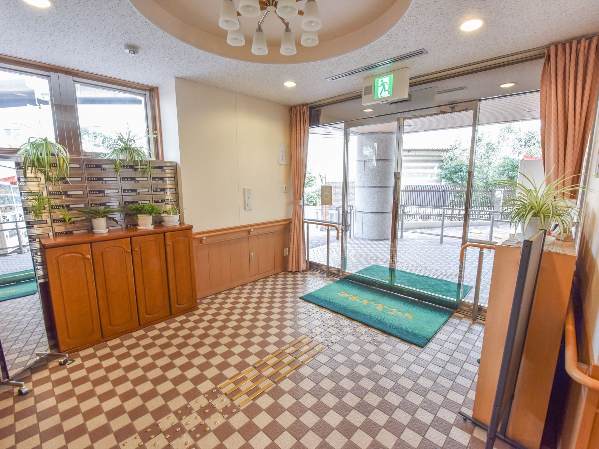 ガラスドアが設置されている明るい雰囲気の玄関。床はブラントとベージュの市松模様になっている。姿見、収納ボックスが置かれている。