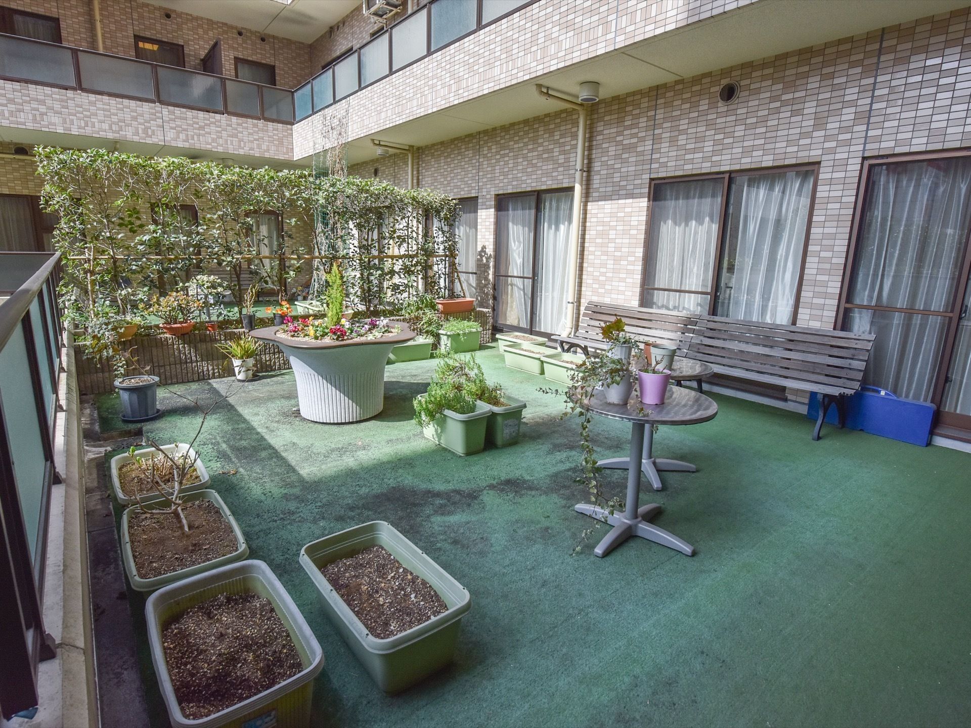陽当たりの良い中庭風のテラスには花の寄せ植えや木々が植えられている。木製のベンチやテーブルが置かれている。