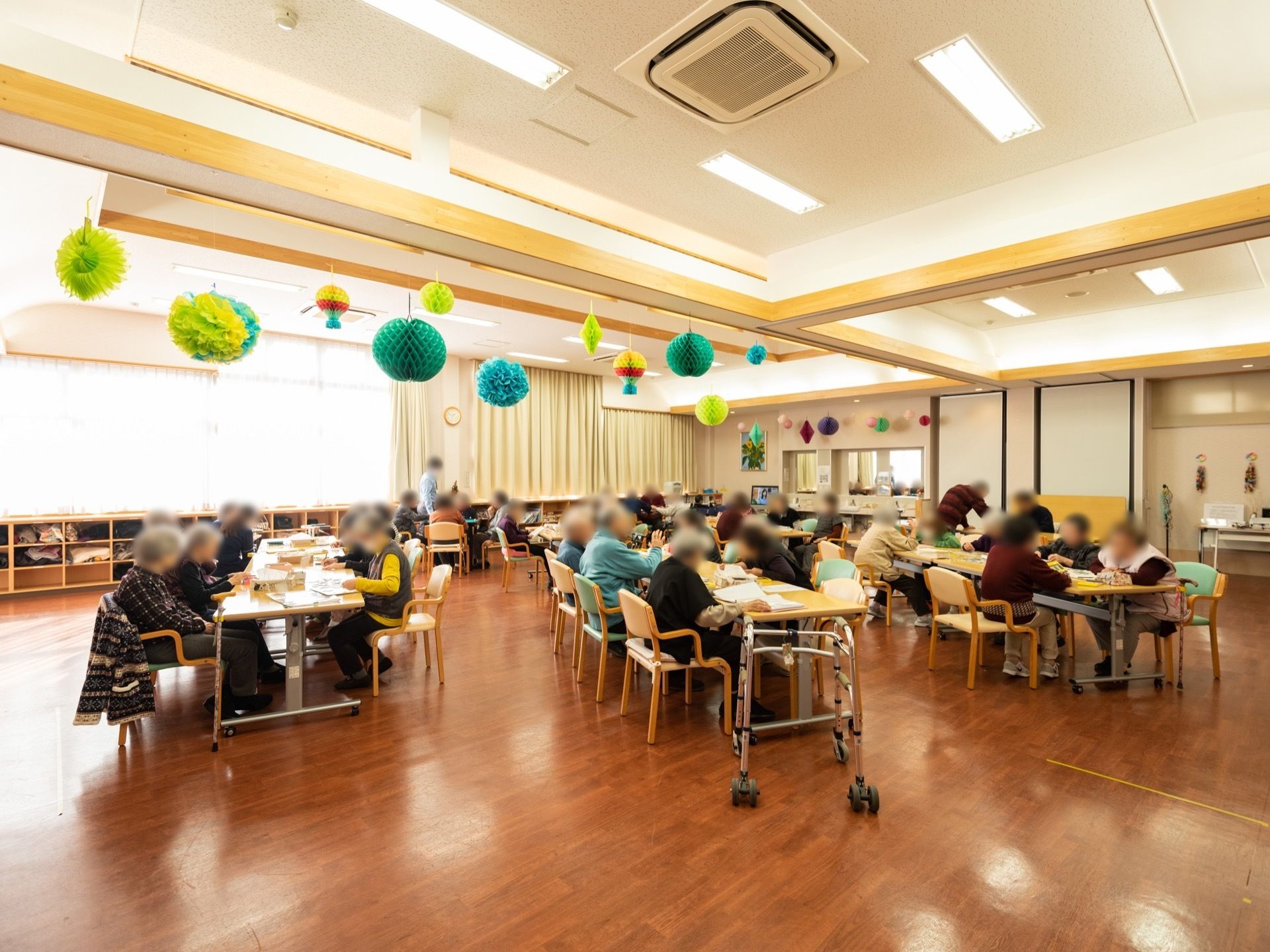 共有スペースで、大勢の入居者様がレクリエーションを楽しんでいる。天井からはたくさんのハニカムボールが下がっている。