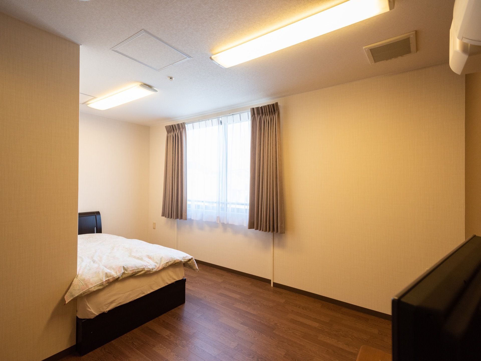 居室は白と茶色を基調にし、落ち着いた雰囲気がある。大きな窓、エアコン、テレビが見える。室内は広々とゆとりがある。