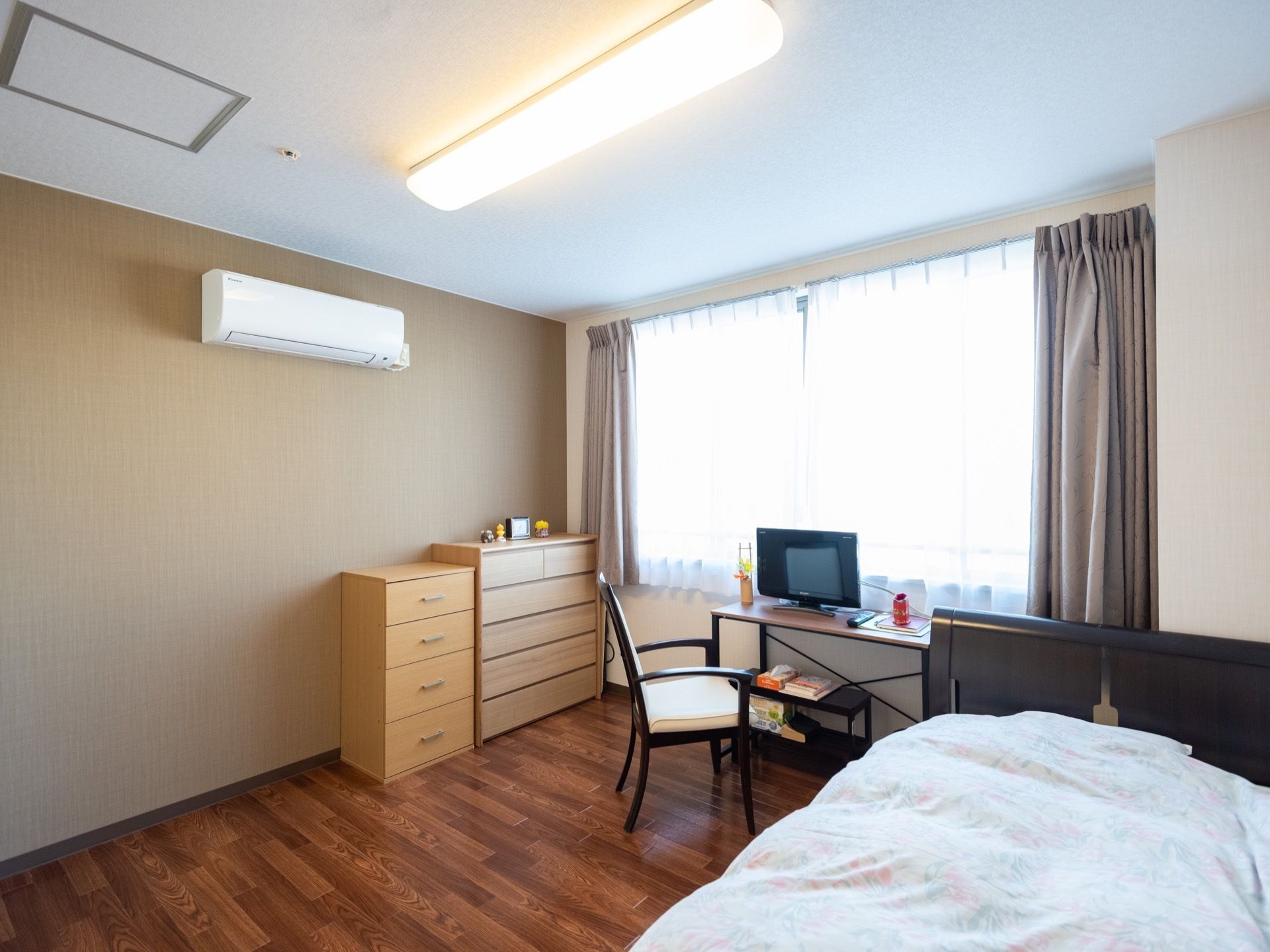 大きな窓があるフローリングの居室。ベッドが１台置かれ、他にもチェスト、洋服ダンス、机などが並べられている。