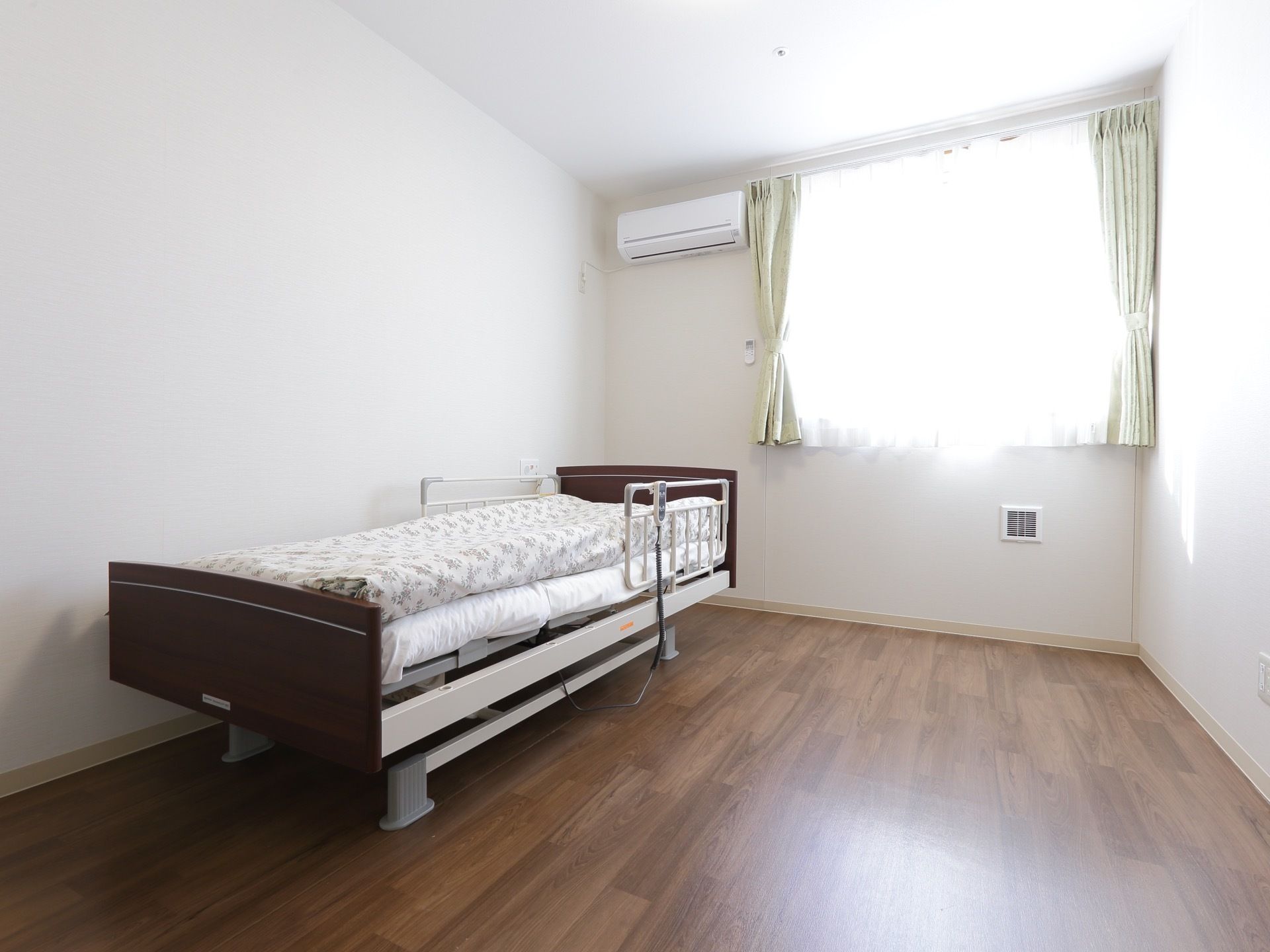 白い壁にダークブラウンのフローリングの部屋である。介護ベッドが置かれている。エアコン、カーテンが備わっている。