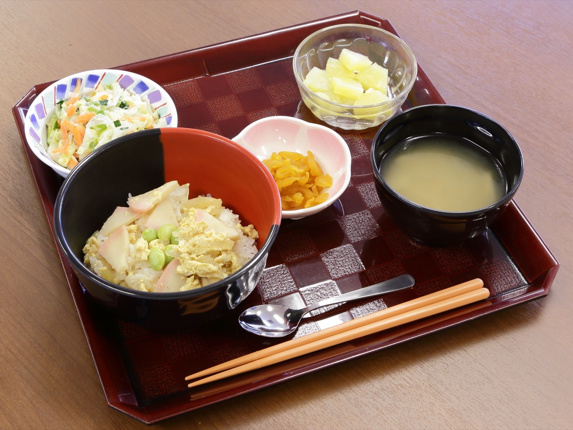 お盆の上に、食事が載っている。かまぼこと枝豆の卵とじご飯、味噌汁、サラダ、漬物、パイナップルのメニューである。