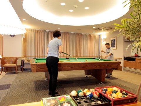 室内の中心にビリヤード台があり、お二人の入居者様がキューを握って対戦しておられる。背後には台所も見える。