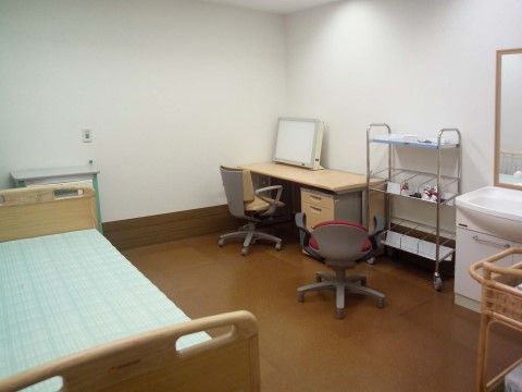 部屋の隅にモニターが載った机と椅子があり、その横には洗面台がある。机の反対側の壁際にはベッドが置いてある。