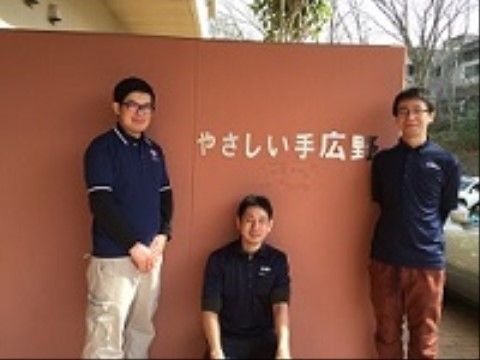 施設名が書かれたブラウンの壁面の前に並び微笑む施設職員