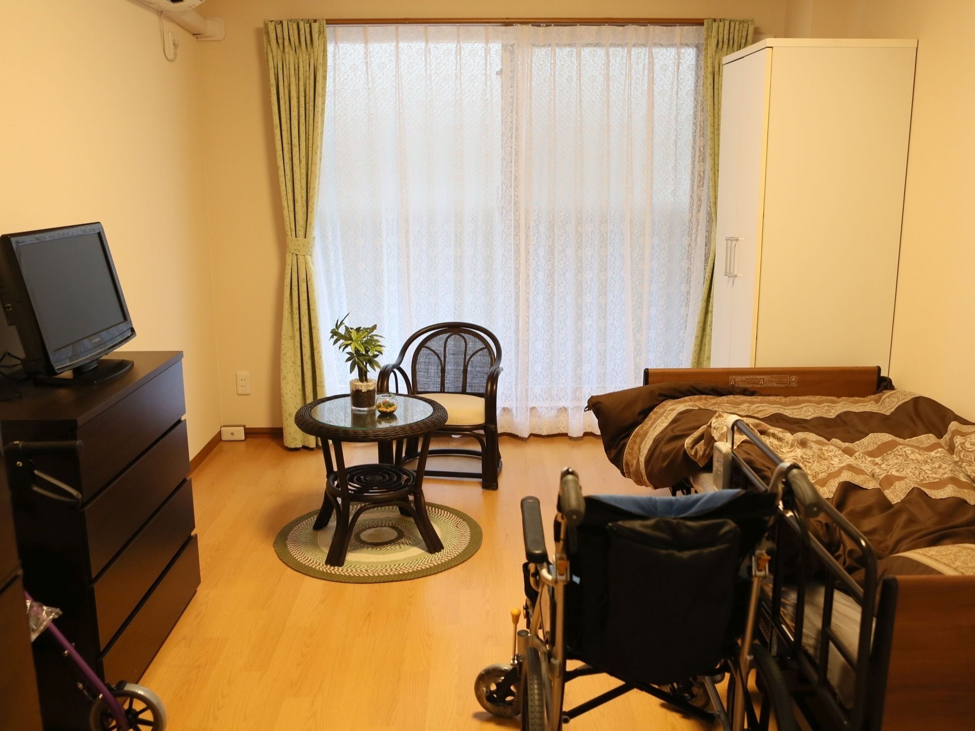 レースのカーテンが掛かった車椅子や落ち着いた色の生活家具がある部屋