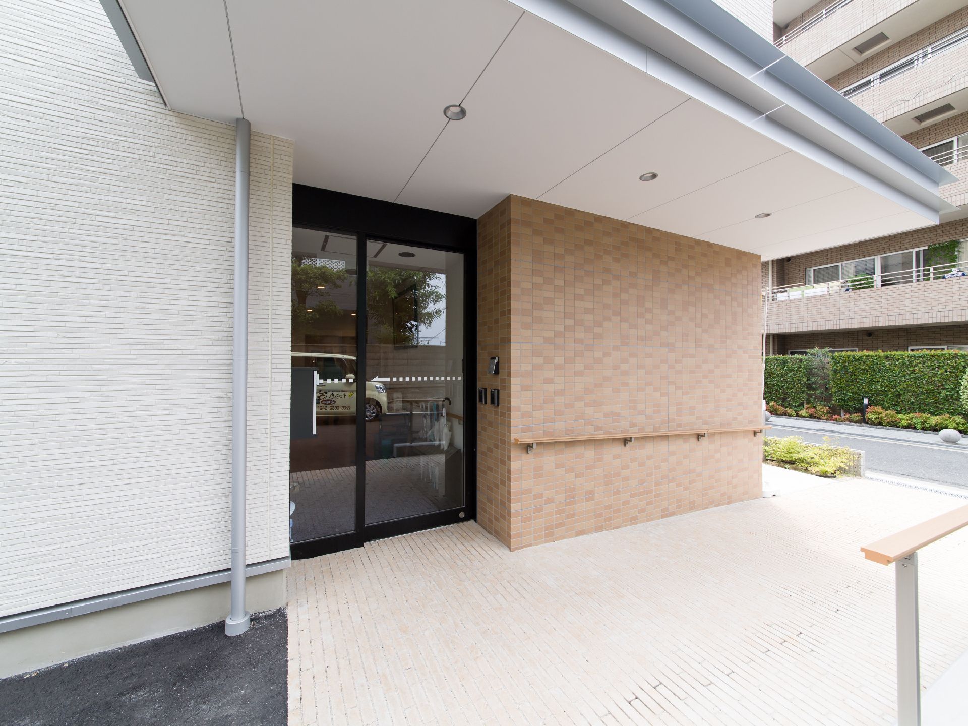 施設の玄関前は明るいベージュのタイル敷きである。上部には大きく屋根がせり出しており、丸い照明が3つ付いている。