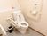 白っぽい壁と白い床に白い便器が置かれたトイレである。便器近くの壁にL字型の手すりがあり、反対側には可動式の手すりが備わっている。
