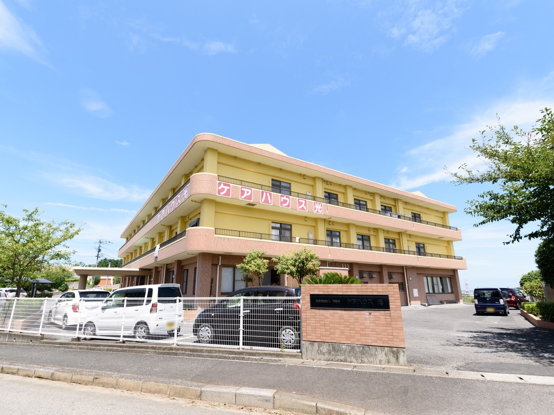 広い敷地の中に3階建ての建物がある。壁はオフホワイトとピンクベージュでまとめられて、優しい雰囲気である。車も多く停まっている。
