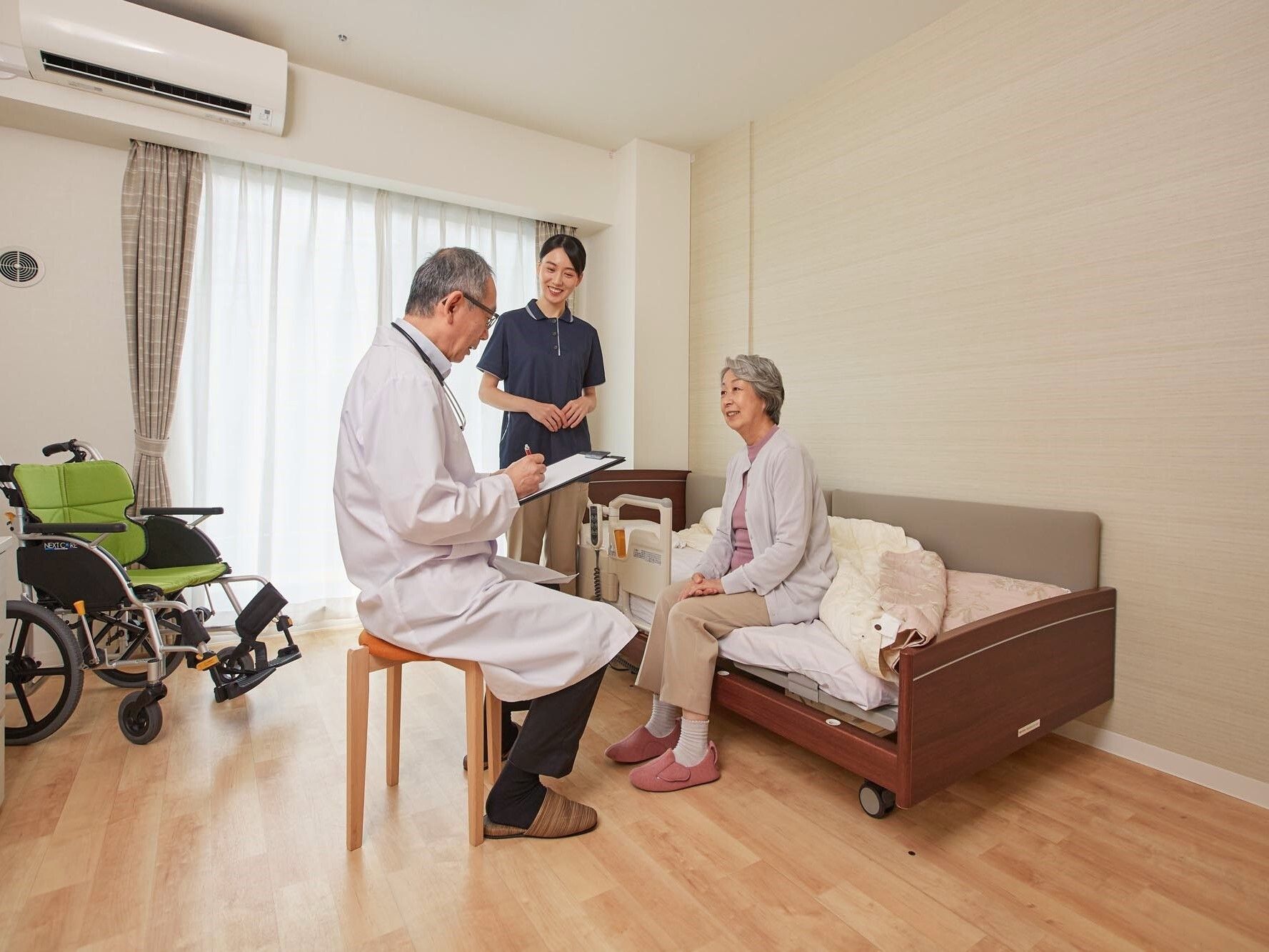 自室のベッドに腰掛け医者と話をする高齢者とそれを見守る職員