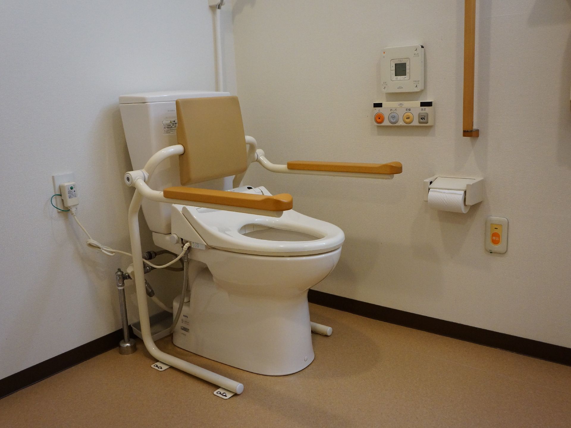 白い壁に茶色の床。便器の両側には手すりがあり、後部には背もたれがある。また、横の壁には縦に手すりが付けられている。