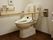 白壁に深みのある焦げ茶色の床のトイレスペース。便器の両側には手すりが設置され、横の壁には流すボタンが取り付けてある。