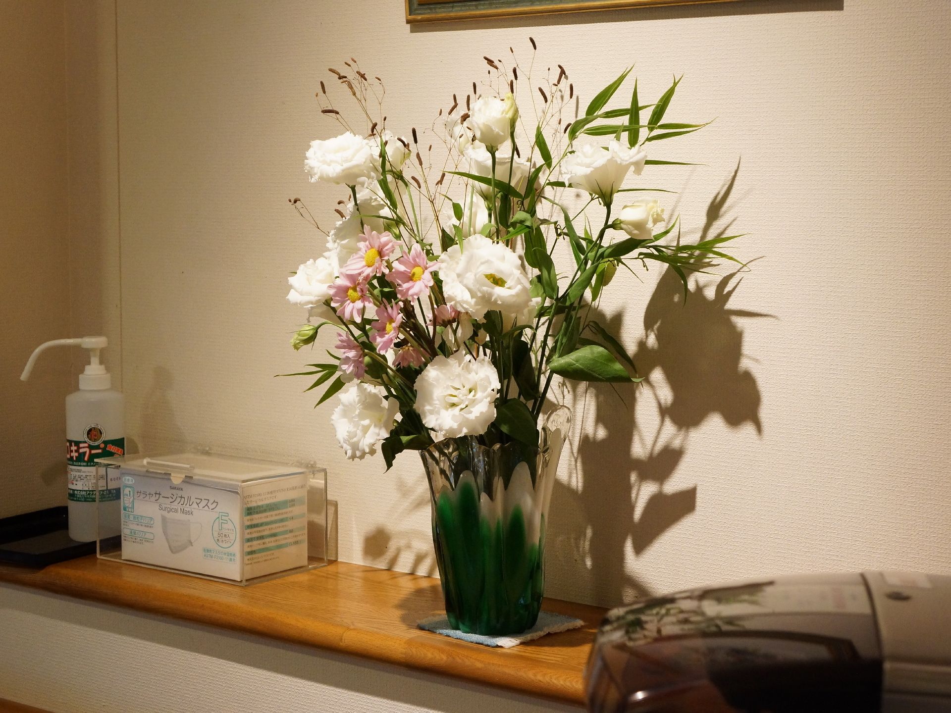 白い壁際に設置された木目調の台の上には花瓶に飾られた花が置かれ、その隣には、マスクや消毒液が置かれている。