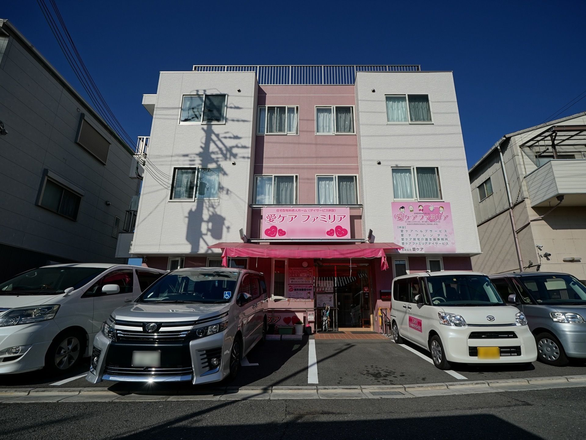 施設の建物は3階建てで、1階の玄関の前には駐車スペースが設けられている。玄関にはピンク色のひさしが備わっている。