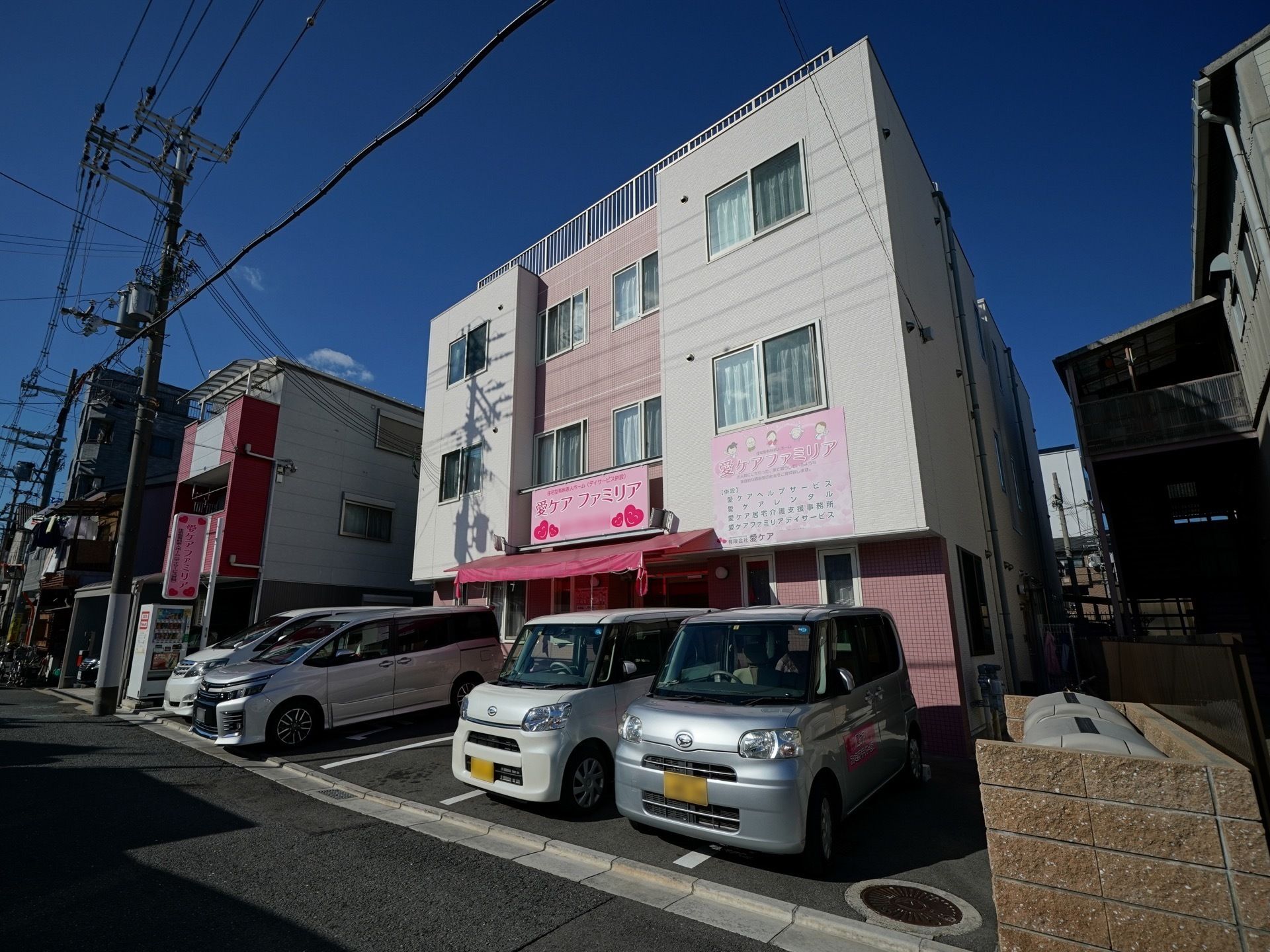 ピンク色を基調とした施設の外観である。施設前には駐車場が用意されており、来訪者様が自動車で訪問できるように配慮されている。