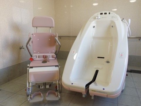 浴室用の車椅子には、シートベルトや足置きが取り付けられている。室内は白を基調とした光沢のある壁に、グレーのタイル張りの床となっている。