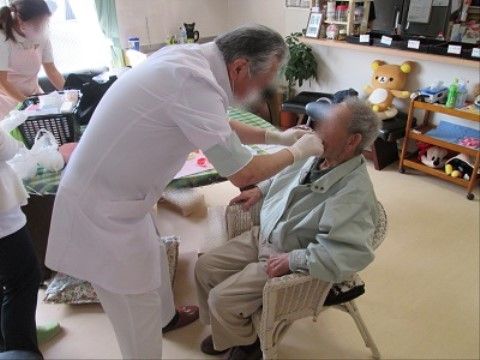 生活雑貨が置かれた部屋で椅子に座り医者に口の中を見てもらう高齢者