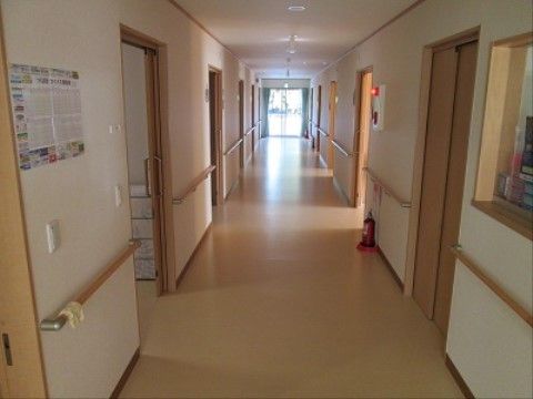 居室の扉がいくつか見える。非常ベルの下に消火器が置かれている。廊下の壁は両側に手すりが設けられている。