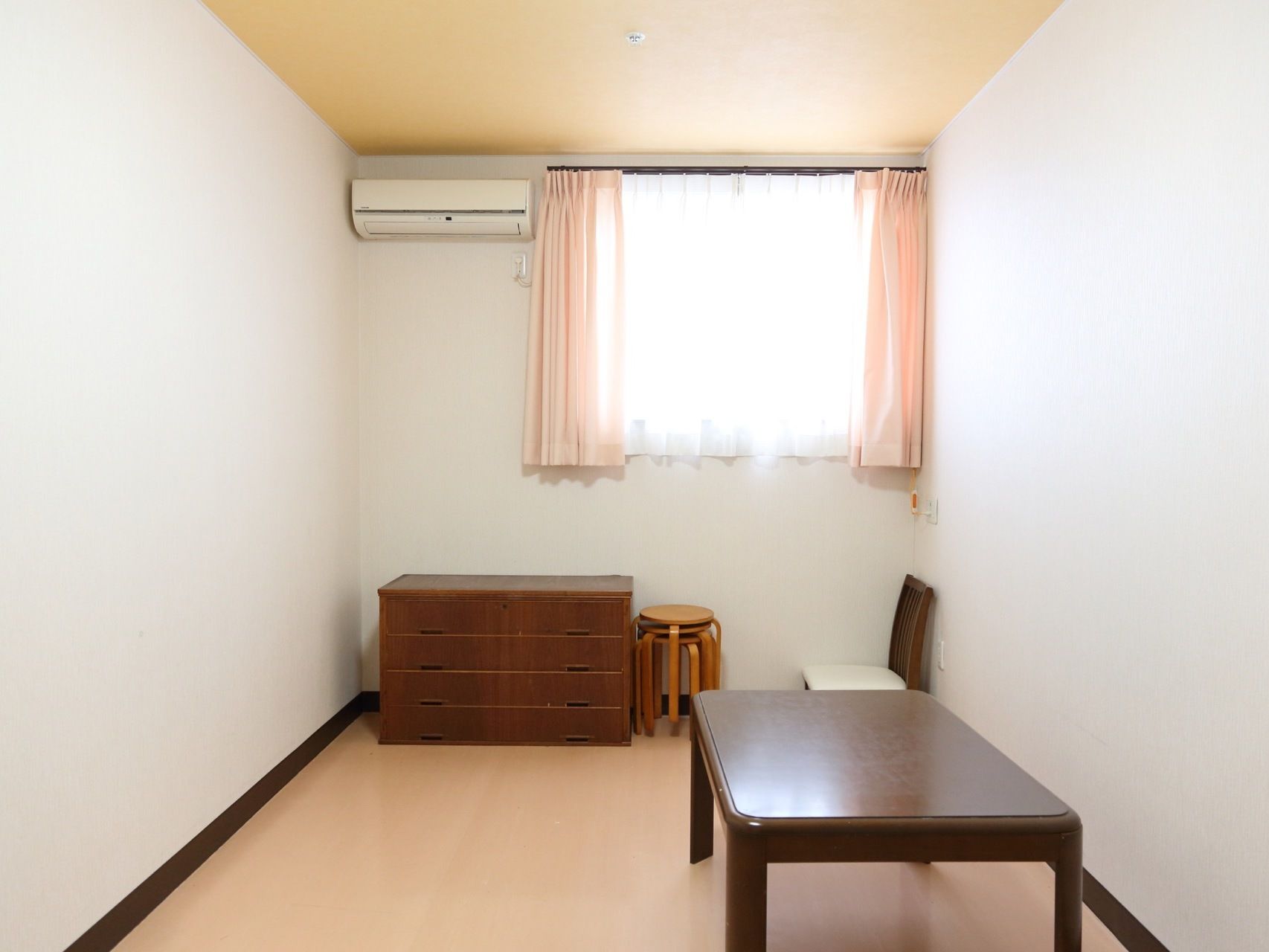 居室は洋室で、エアコンが設置されている。焦げ茶色のチェストとテーブルが置かれている。窓は腰高で、ベージュ系のカーテンが取り付けられている。