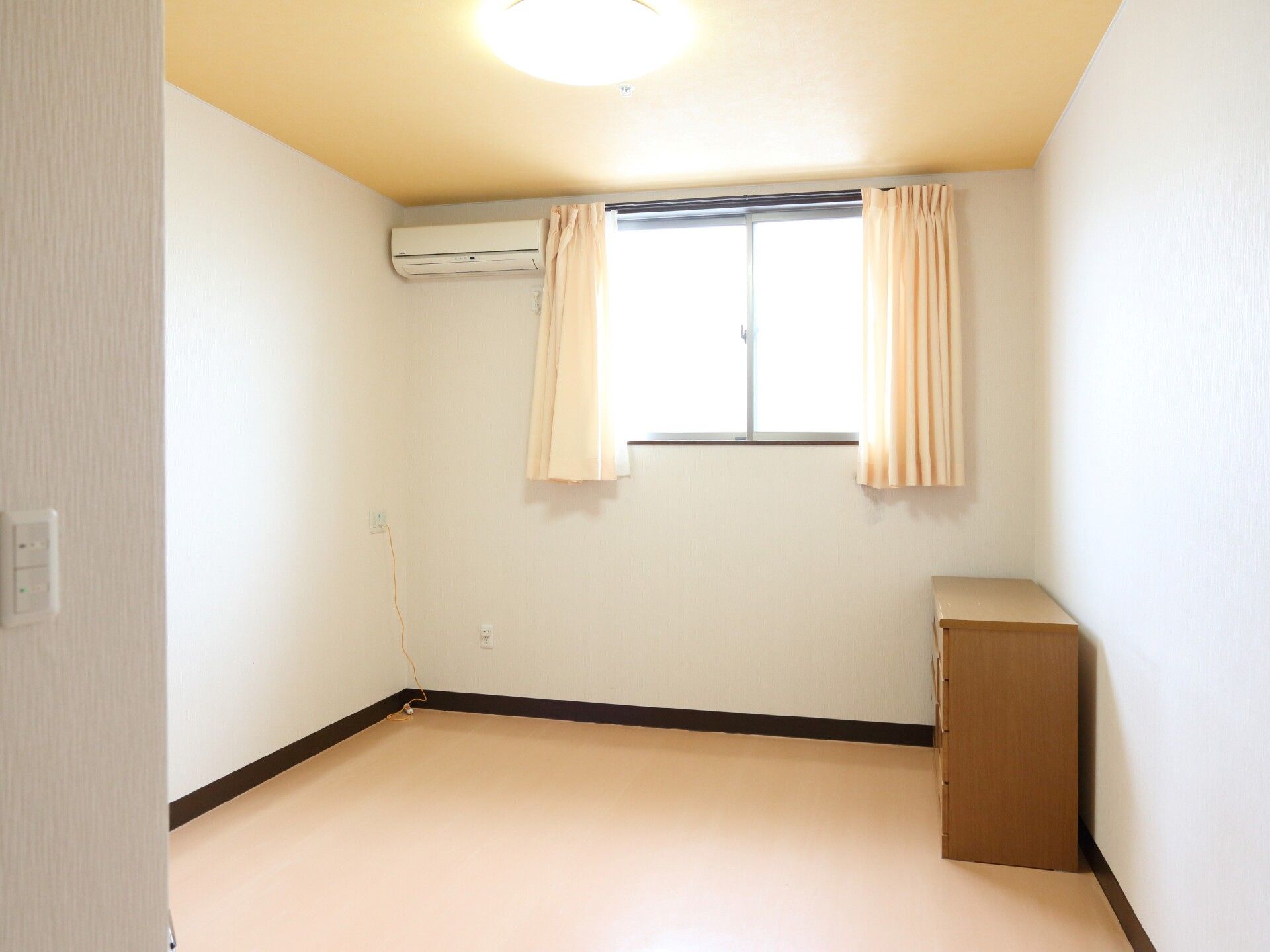 エアコンとチェストが置かれ、窓から日の光が入る白壁の部屋