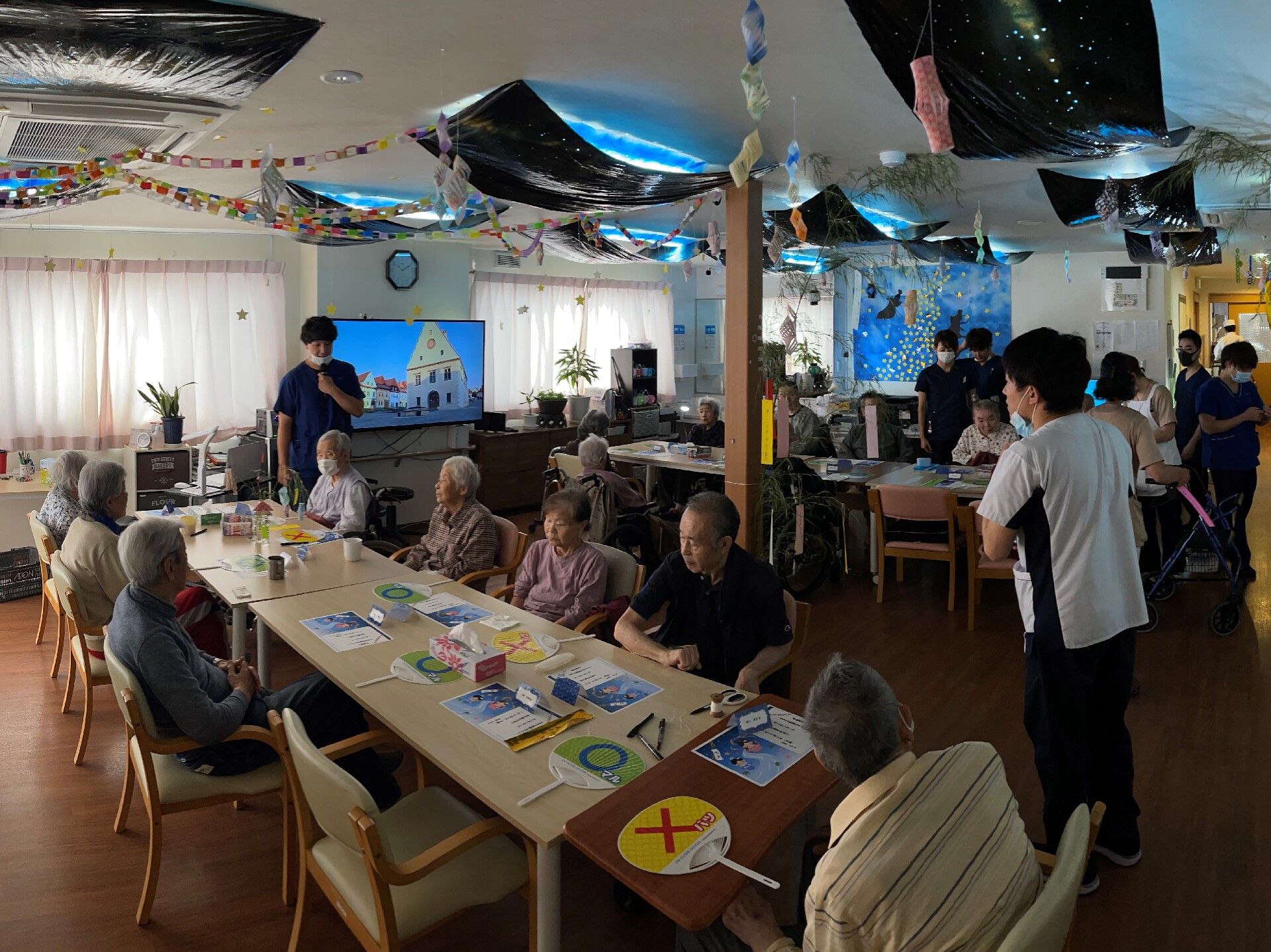 広々とした共有スペースで、入居者様が製作を楽しんでいる。壁側にはテレビが設置されており、植物が置かれている。