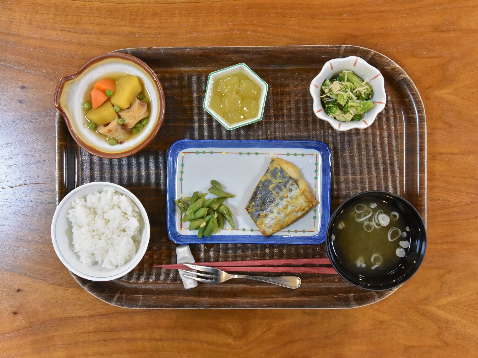 トレーに食事が載っている。ご飯、味噌汁、メインのおかずの皿の他に、複数の小鉢があり、品数が多い。箸と小さなフォークが添えられている。