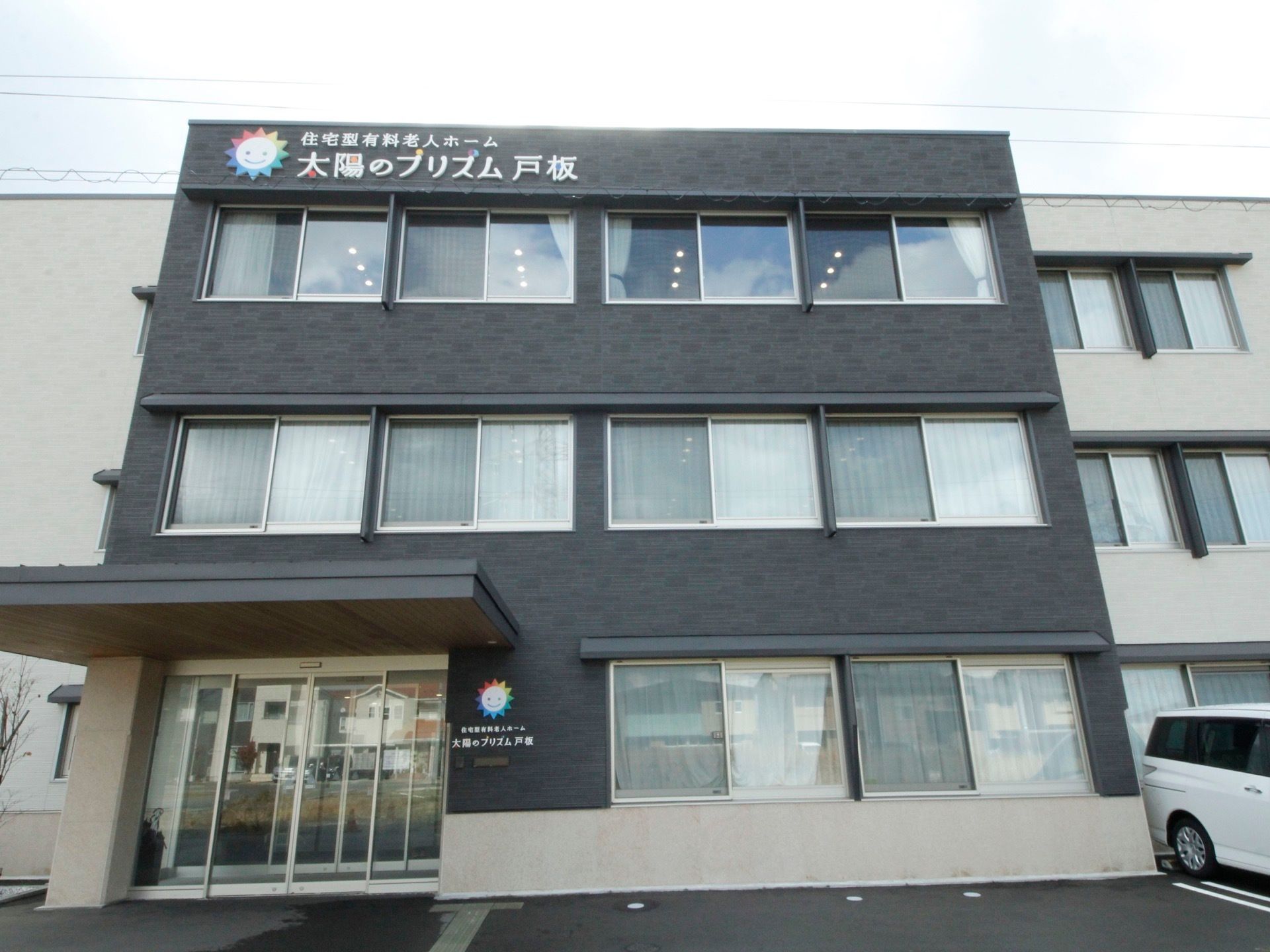 施設名を外壁と玄関わきに大きな文字で表示しており、わかりやすくなっている。黒と薄いベージュの外壁で、落ち着いた雰囲気になっている。