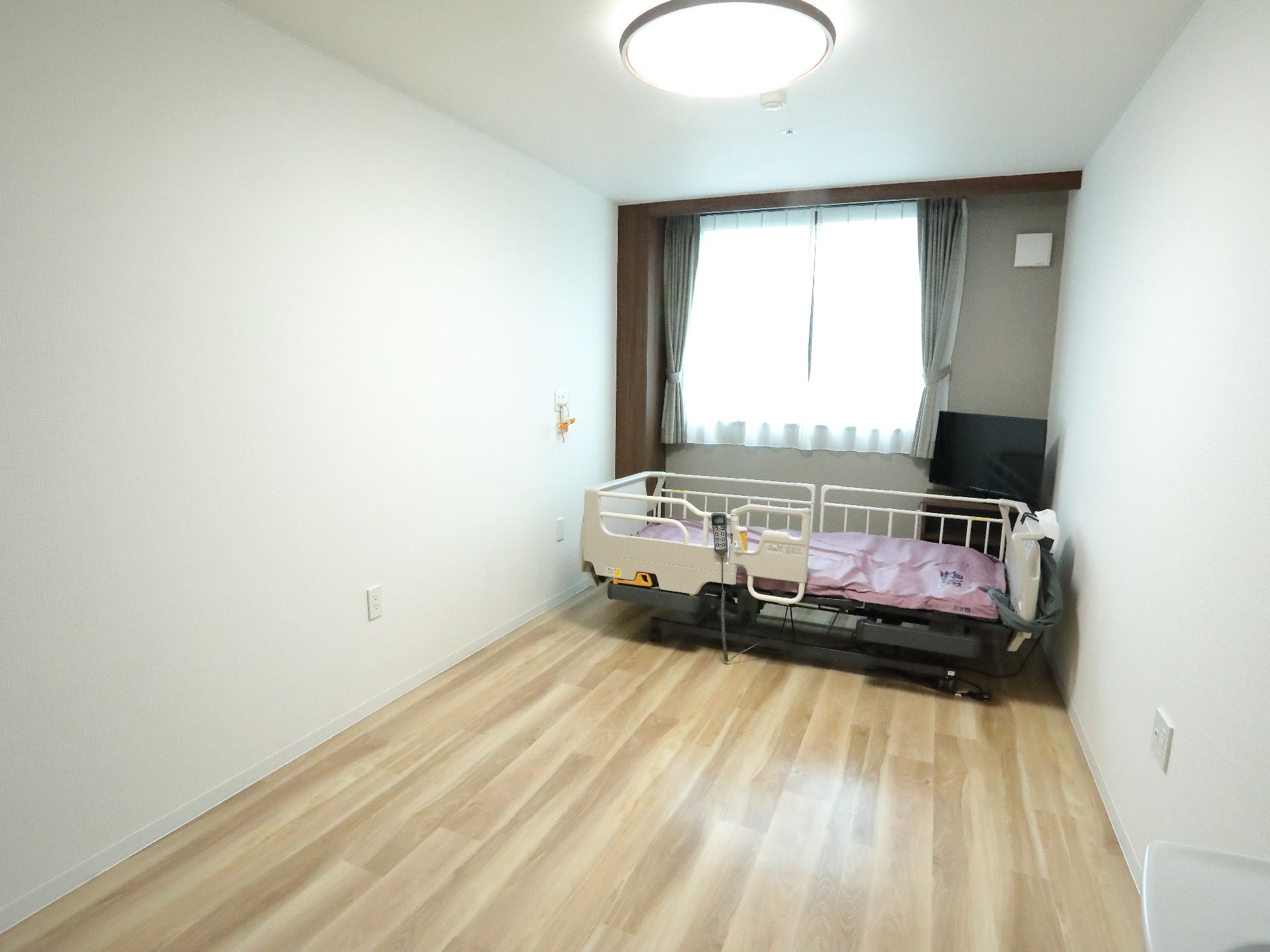 居室の天井には、丸型の大きなライトがある。部屋の奥にはベッドがあり、そのそばにはテレビが置かれている。