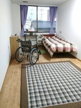 正面に窓がある明るい居室。ベッドカバー、車椅子、ラグがチェック柄でまとめられていて、ポップな印象の部屋になっている。