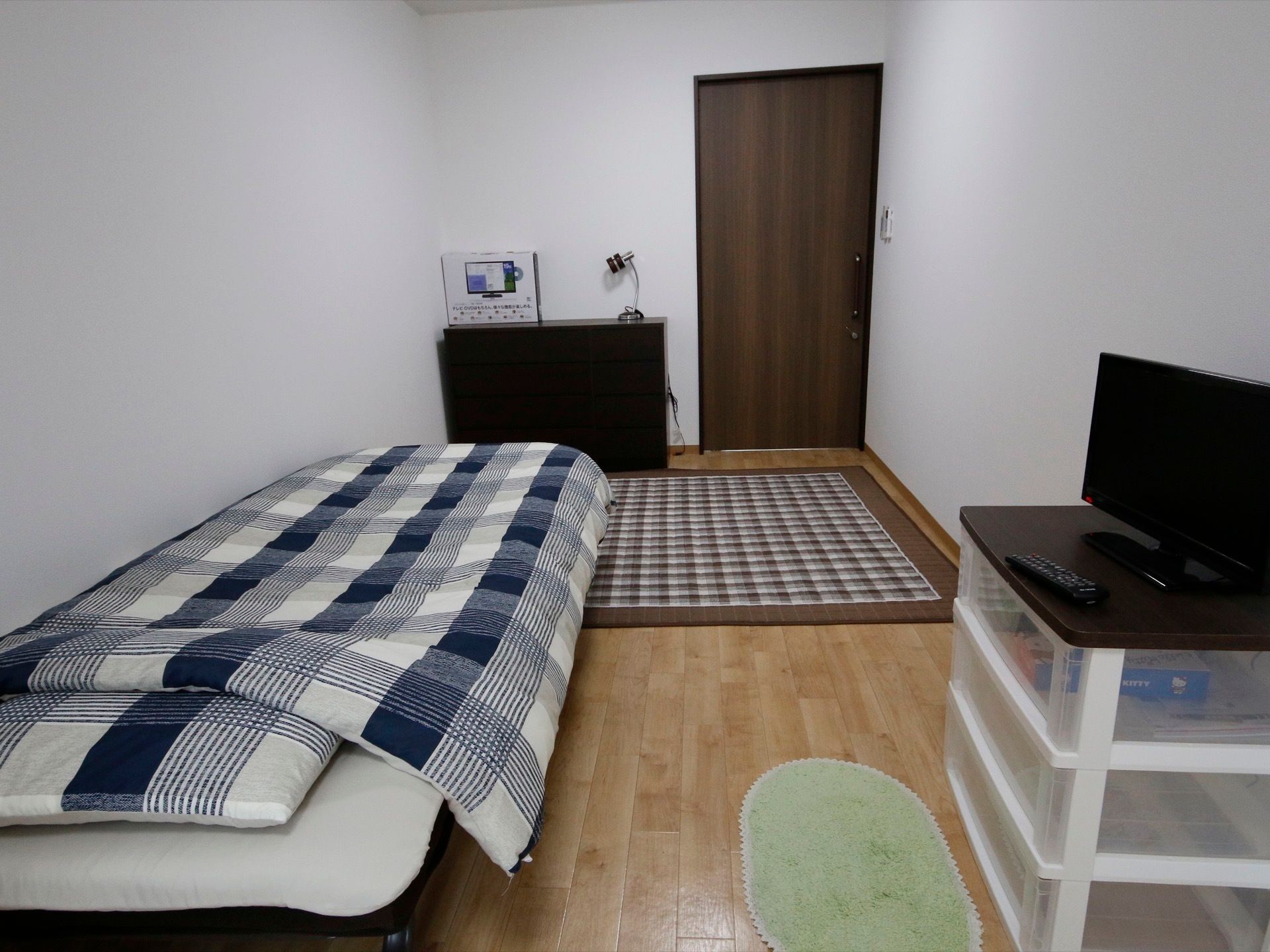 白い個人部屋にベッド、カーペット、小さな箪笥、チェストがある。タンスの上にライト、チェストの上にテレビが置かれている。